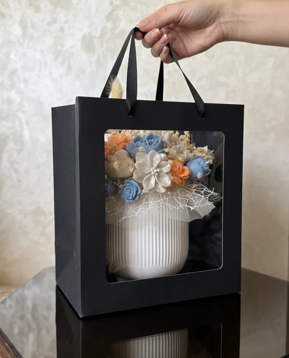 Le bouquet corail & bleu dans sa verrine blanche, présenté dans un sac cadeau noir à fenêtre.