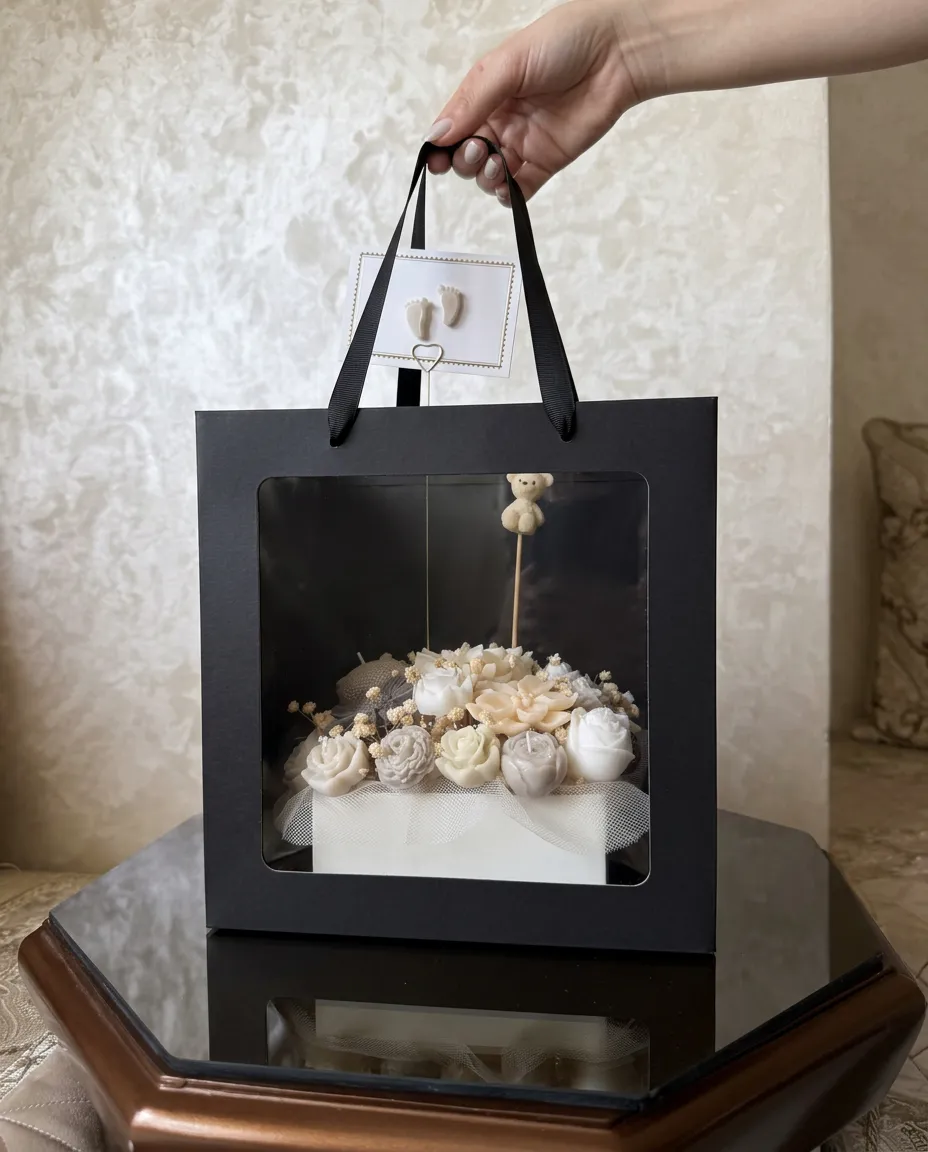Box naissance : roses et fleurs en cire ivoire avec ourson et carte petits pieds, dans un sac cadeau noir à fenêtre.