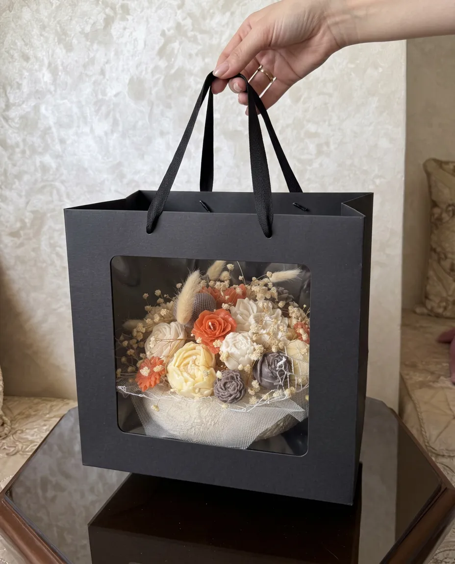 Composition de roses en cire corail, ivoire et taupe dans une coupe blanche, présentée dans un sac cadeau noir.